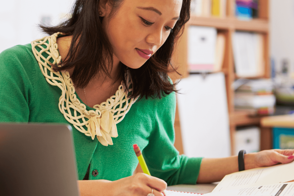 A woman in a green sweater with a decorative collar writes in a notebook with a pencil, seated at a desk with a laptop in a classroom